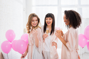 happy multiethnic girls having fun with champagne and cupcake during bachelorette party with pink balloons