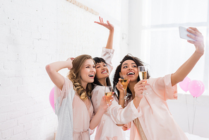 excited multicultural girls with glasses of champagne taking selfie on smartphone during pajama party