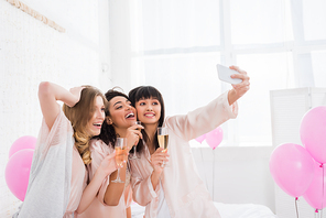 attractive multicultural girls with glasses of champagne taking selfie on smartphone during pajama party