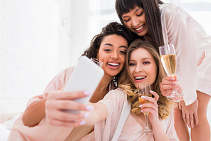 smiling multiethnic girls with champagne glasses taking selfie on smartphone during pajama party