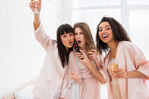 excited multiethnic girls holding champagne and singing with microphone on pajama party