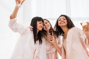 beautiful happy multicultural girls holding champagne and singing with microphone on pajama party