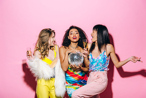 cheerful multiethnic girs with champagne and disco ball dancing on pink