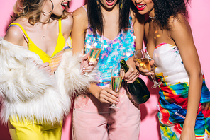 cropped view of emotional multicultural girlfriends pouring champagne from bottle into glasses on pink
