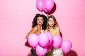 multicultural smiling girlfriends holding milkshakes on pink with balloons