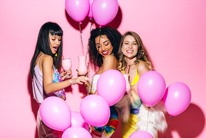 cheerful stylish multiethnic girlfriends holding milkshakes on pink with balloons
