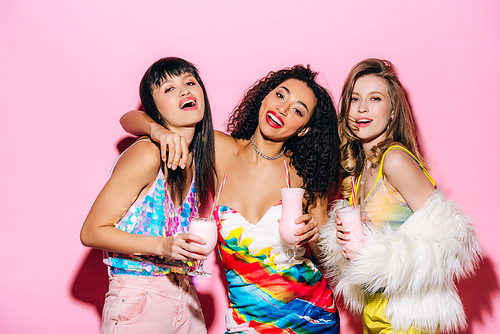 smiling stylish multiethnic girls holding glasses with milkshakes on pink
