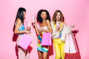 smiling stylish multiethnic girlfriends posing with milkshakes and shopping bags on pink