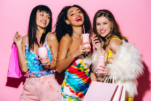 excited fashionable multiethnic girls posing with milkshakes and shopping bags on pink