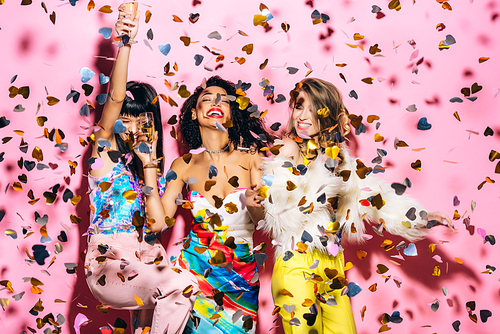 happy multicultural girls having fun with glasses of champagne on pink with confetti