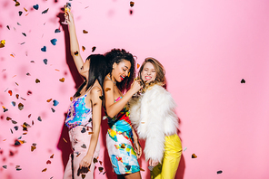 excited multicultural girls dancing with glasses of champagne on pink with confetti