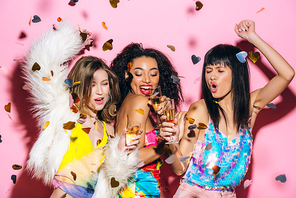 excited fashionable multicultural girls dancing with glasses of champagne on pink with confetti