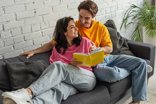 smiling young couple resting on couch, hugging and holding book in living room