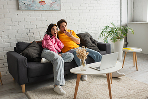 amazed young couple sitting on couch with open mouths and watching movie on laptop in living room