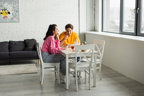 positive young couple sitting at table near window and eating breakfast in modern living room