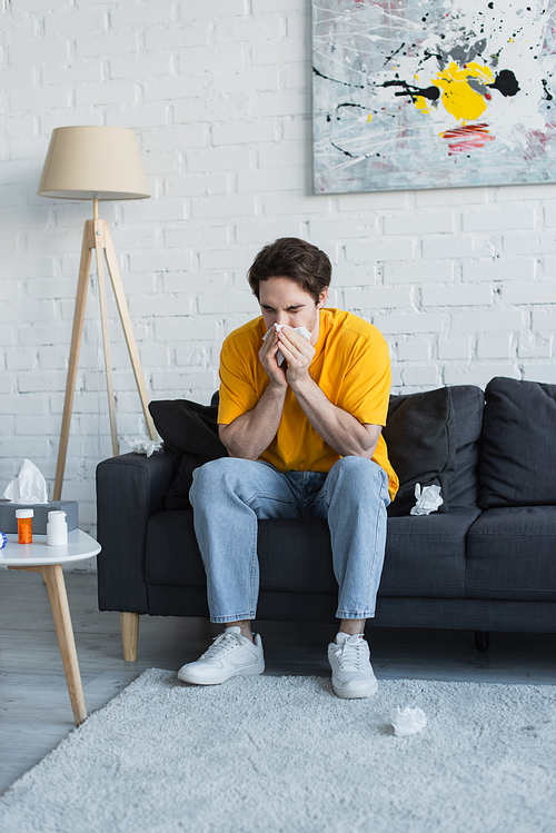 diseased young man sitting on couch and blowing nose with paper napkin at home