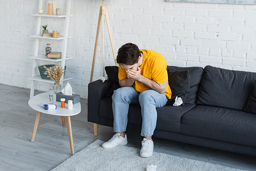 sick young man sitting on couch and sneezing with hands covering face at home