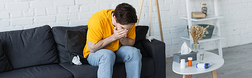 diseased young man sitting on couch and sneezing with hands covering face at home, banner