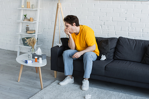 sick young man sitting on couch and coughing with hand near face at home