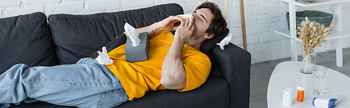 sick young man lying on couch and blowing nose with paper napkin at home, banner
