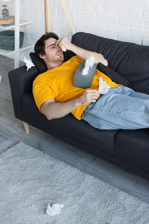 diseased young man lying on couch with hand near head and holding paper napkin at home