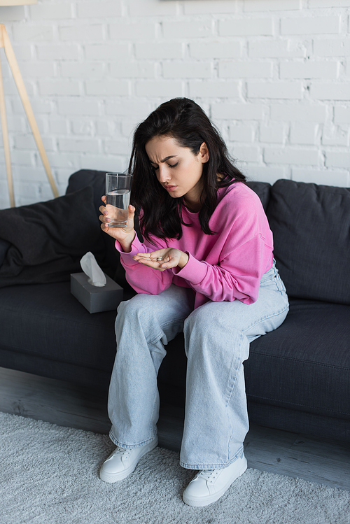 diseased young woman sitting on couch with glass of water and looking at pills in palm in living room