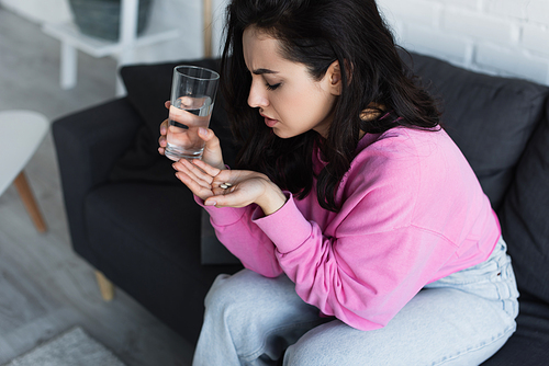 diseased young woman sitting on couch with glass of water and pills in living room