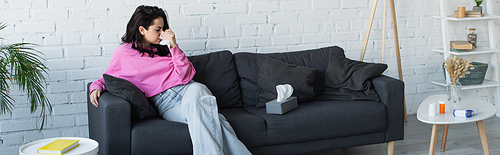 diseased young woman sitting on couch and blowing nose with paper napkin in living room, banner