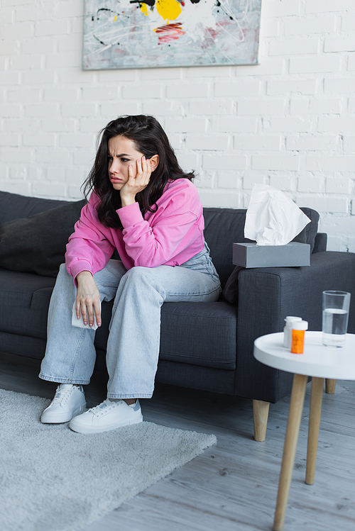 sick young woman sitting on couch with hand near face and holding paper napkin in living room