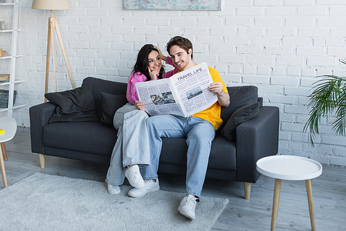 smiling young couple sitting on couch and holding newspaper at home