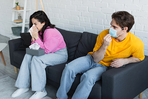 diseased young woman sitting on couch and blowing nose with paper napkin near boyfriend adjusting medical mask at home