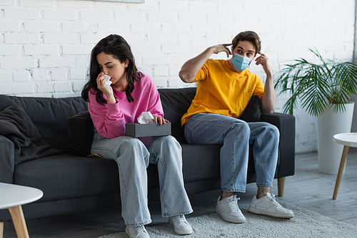 diseased young woman blowing nose with paper napkin near boyfriend sitting on couch and adjusting medical mask at home