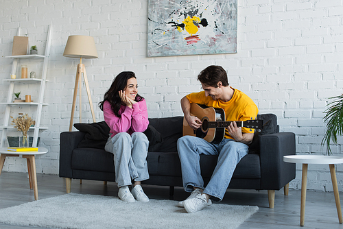 young man playing acoustic guitar near girlfriend with hands near face at home