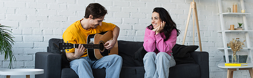 young man playing acoustic guitar near girlfriend with hands near face at home, banner