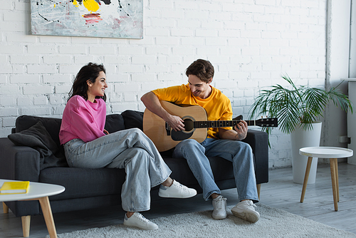 smiling young man playing acoustic guitar near girlfriend sitting on couch at home