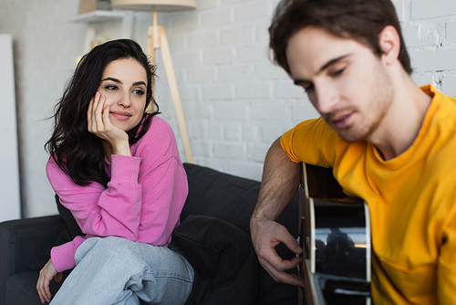 smiling young woman sitting on couch with hand near face near blurred boyfriend playing acoustic guitar at home