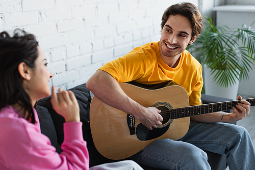 smiling young man sitting on couch with acoustic guitar near girlfriend at home