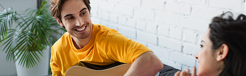 smiling young man sitting on couch with acoustic guitar near girlfriend at home, banner