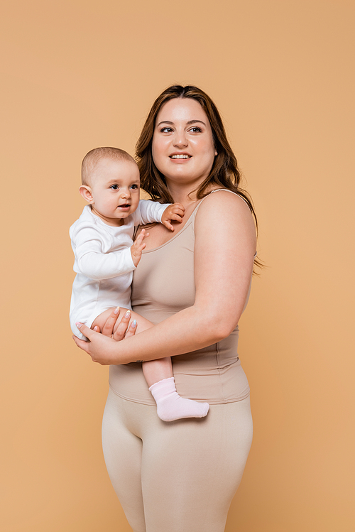 Happy woman with overweight holding baby and looking away isolated on beige