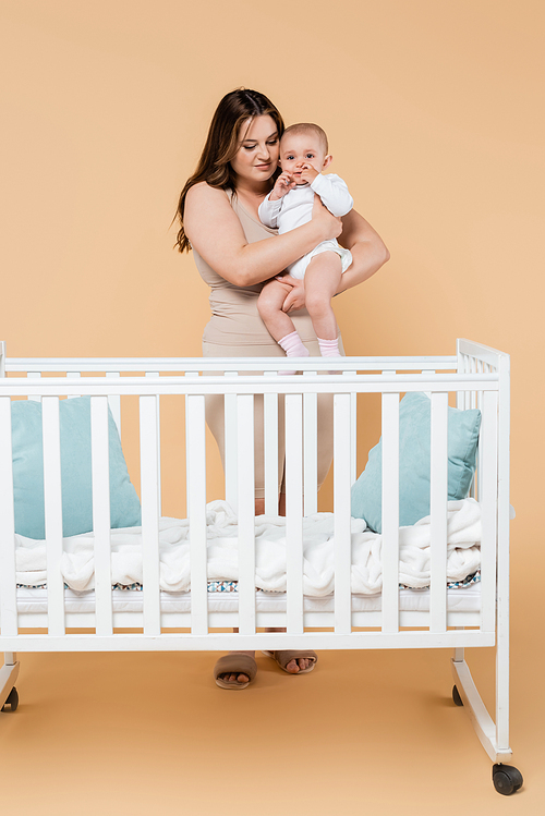 Young plus size woman holding baby near crib on beige background
