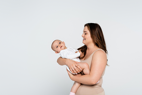 Cheerful brunette plus size mother looking at baby girl isolated on grey