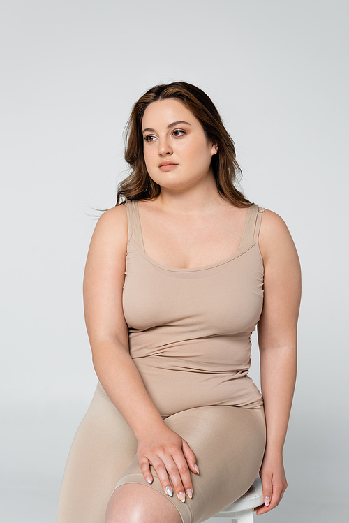 Pretty body positive woman looking away while sitting on chair isolated on grey
