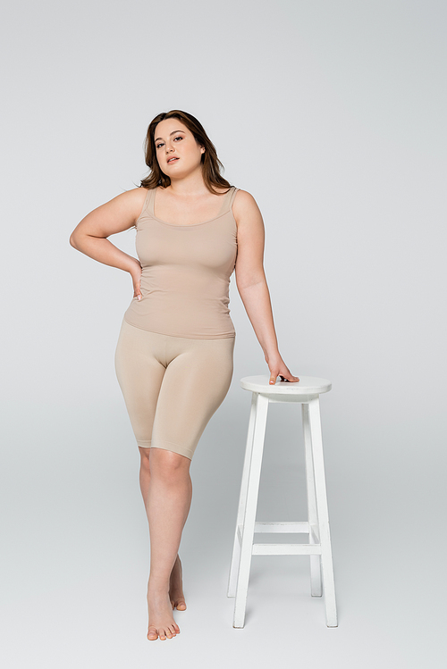 Full length of pretty plus size woman holding hand on hip near chair on grey background