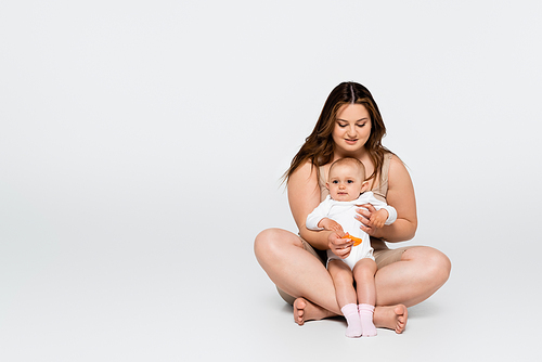 Cheerful body positive woman hugging baby daughter while sitting on grey background
