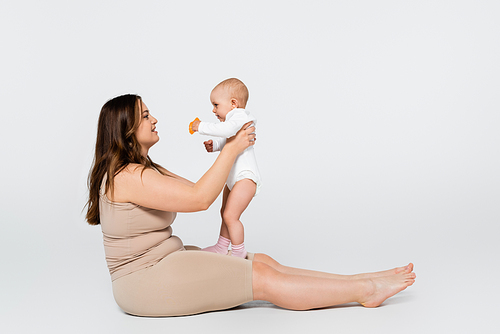 Side view of happy plus size mother holding baby with toy on grey background