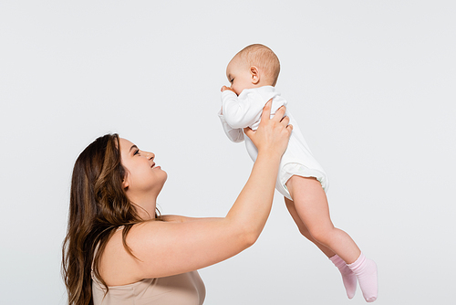 Side view of happy plus size mother lifting baby isolated on grey