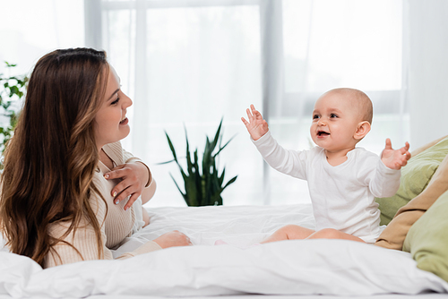 Cheerful baby daughter sitting near brunette mother on bed at home