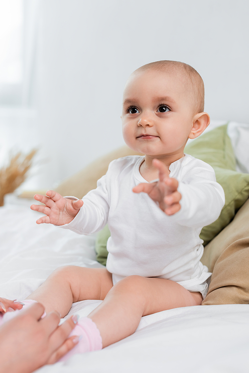 Baby girl sitting on bed near blurred hands of mother