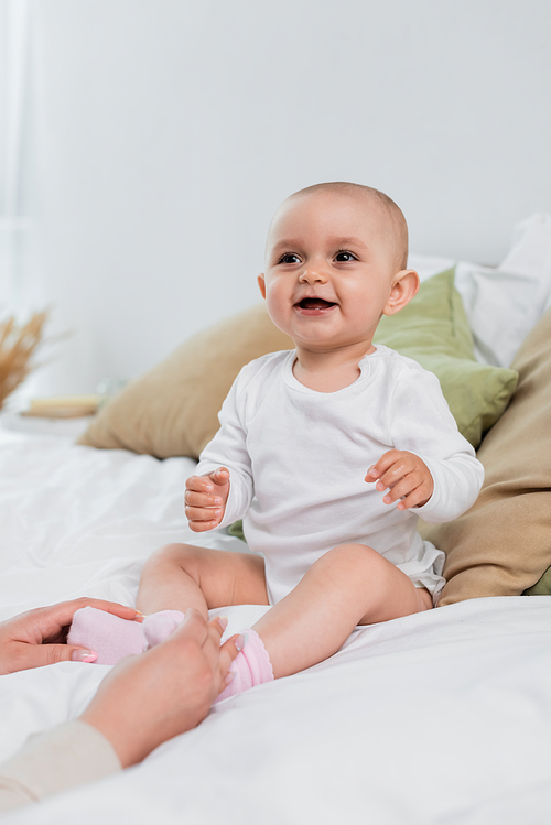 Cheerful baby girl sitting on bed near hands of mom