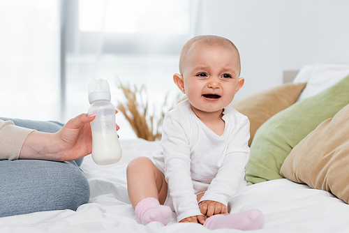 Woman holding baby bottle with milk near cheerful daughter on bed